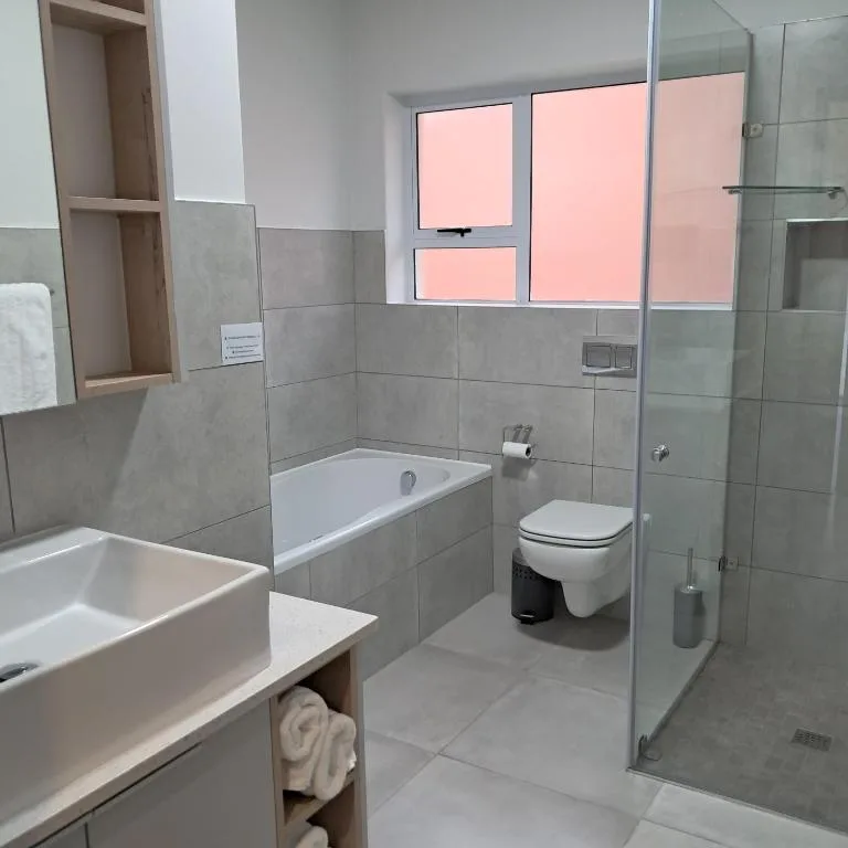 Modern bathroom with grey tile walls, white bathtub, toilet, and glass shower enclosure