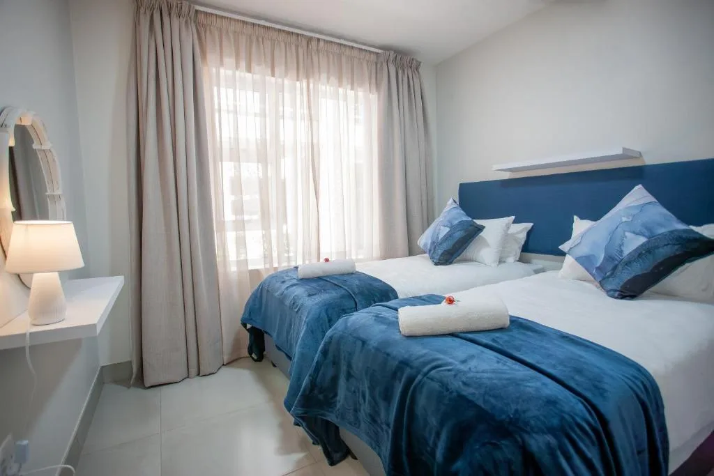 Modern bedroom with blue headboard, twin beds, and sheer curtains allowing natural light