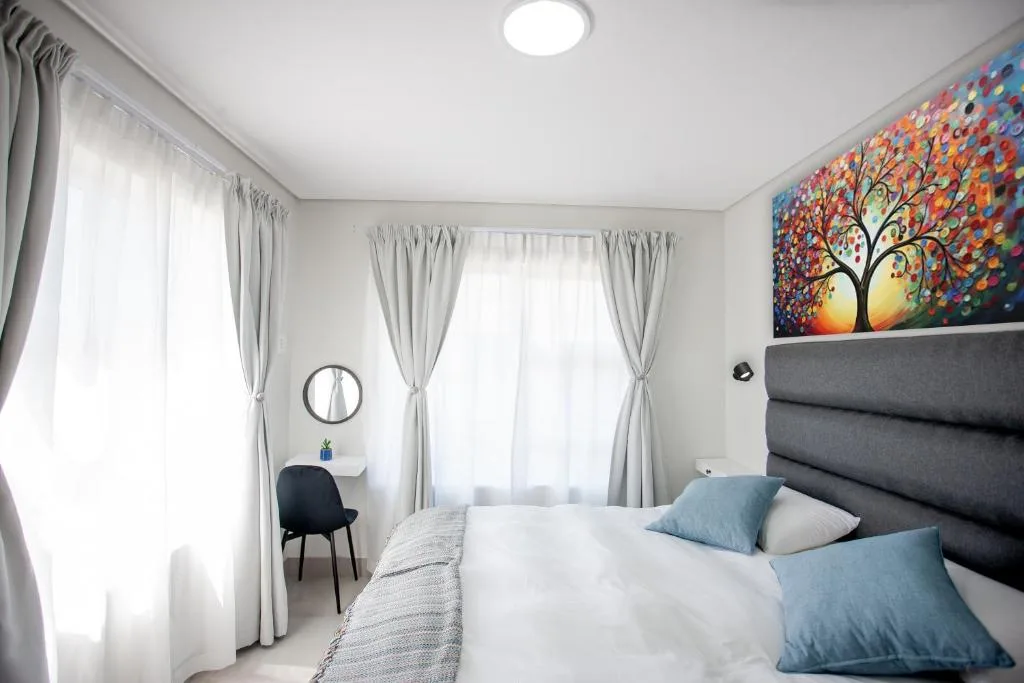 Bright bedroom with queen bed, grey headboard, colourful artwork, and large windows