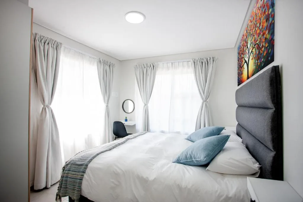 Bright bedroom with queen bed, gray headboard, and multiple windows with sheer curtains