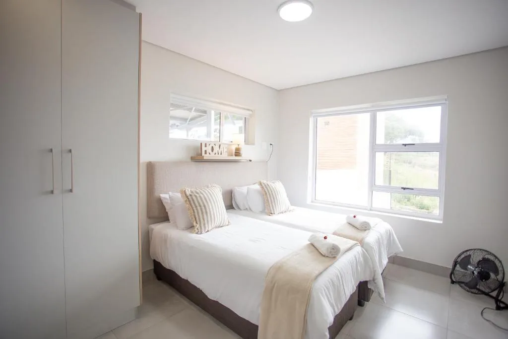 Bright bedroom with twin beds, striped pillows, and large windows overlooking gardens