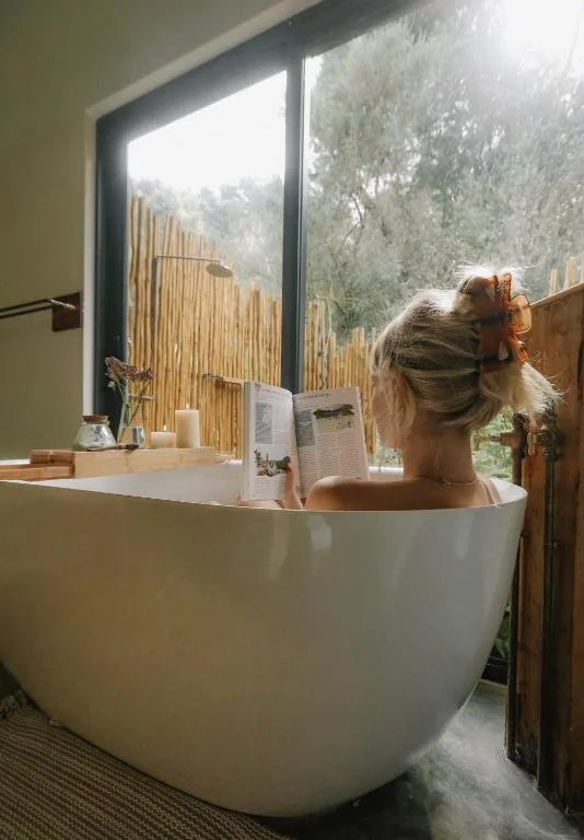 Woman relaxing in modern white freestanding bathtub with scenic garden view through large window