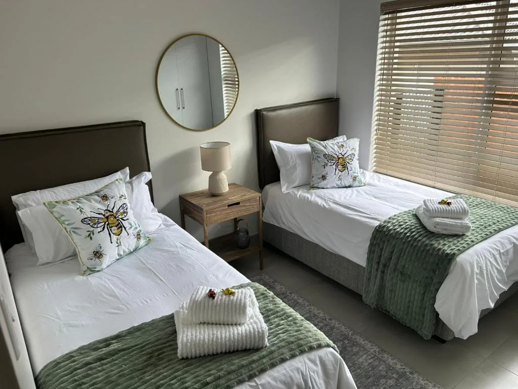 Twin bedroom with white linens, bee-themed pillows, green throws, and round mirror