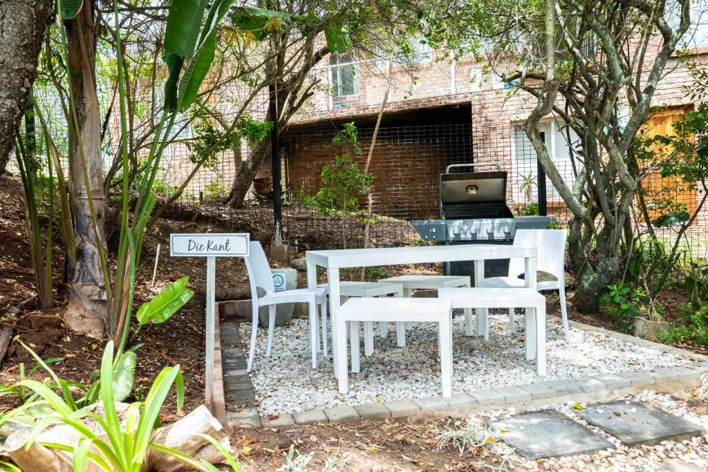 Shaded outdoor braai area with white table and benches surrounded by trees