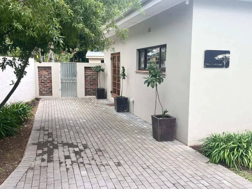 Modern white cottage entrance with paved driveway, potted plants, and brick gate
