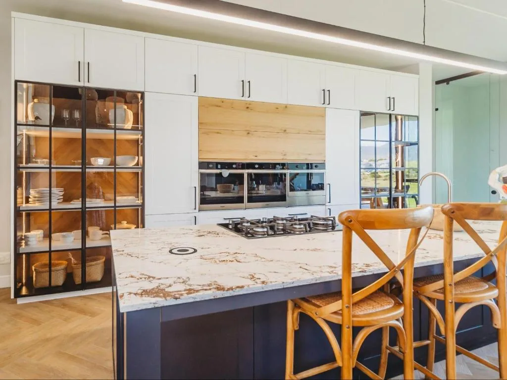 Modern kitchen with marble island, built-in ovens, gas cooktop, and wooden chairs
