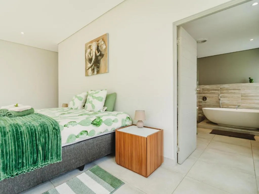 Bright bedroom with green leaf bedding, wooden nightstand, and ensuite bathroom visible