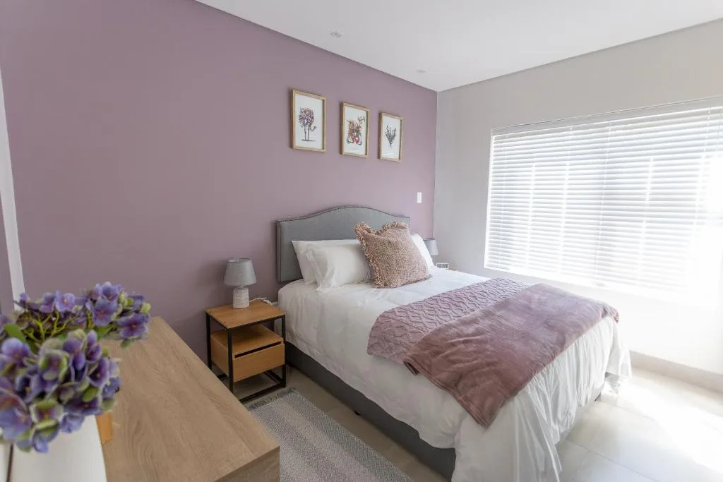 Bright bedroom with purple accent wall, white bed, and botanical prints