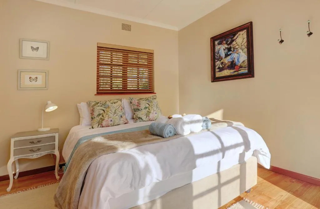 Bright bedroom with neatly made bed, floral pillows, wooden shutters, and artwork