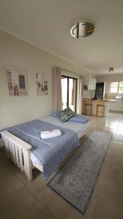 Bright bedroom with blue bedding, white frame bed, and open-plan kitchen visible beyond