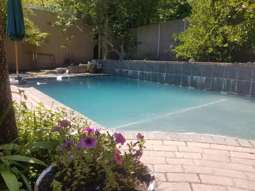 Turquoise swimming pool with brick paving, flowering plants, and shaded trees