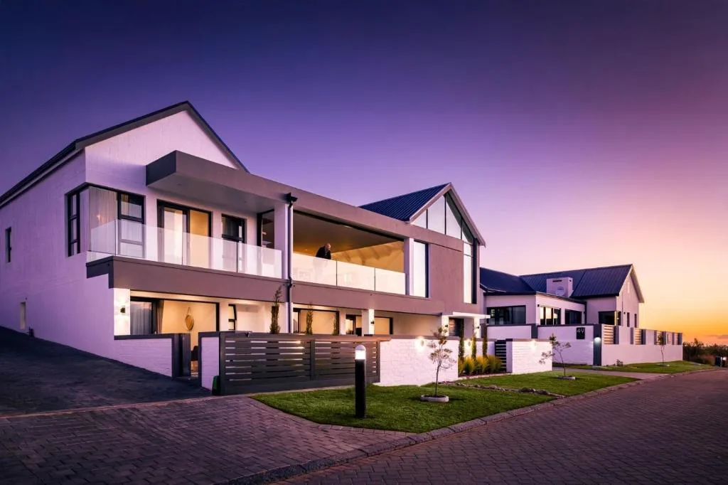 Modern white and brown apartment building at dusk with illuminated windows and manicured lawn