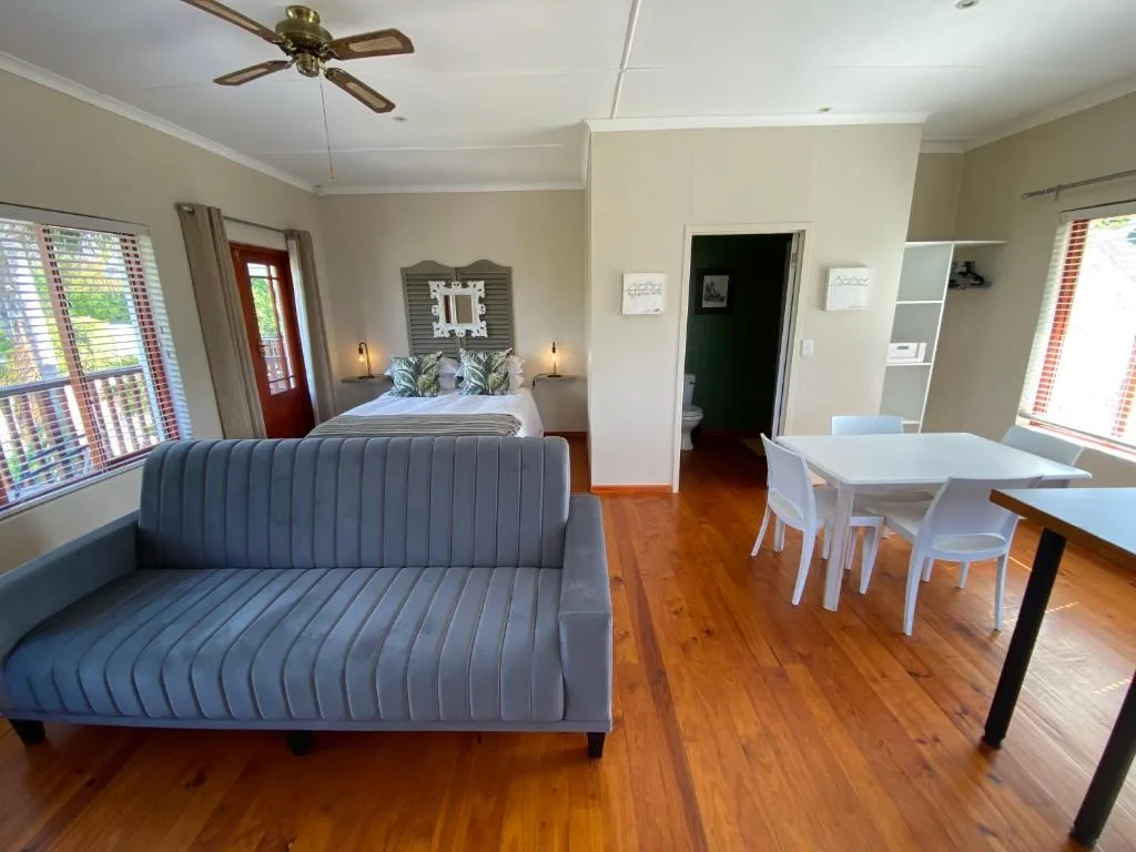 Open-plan lounge with blue sofa, wooden floors, ceiling fan, and bedroom beyond