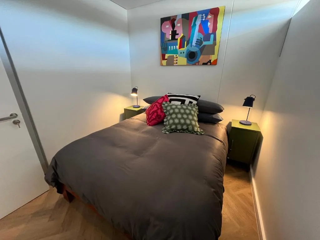 Modern bedroom with dark bed, colorful artwork, bedside lamps, and wooden flooring
