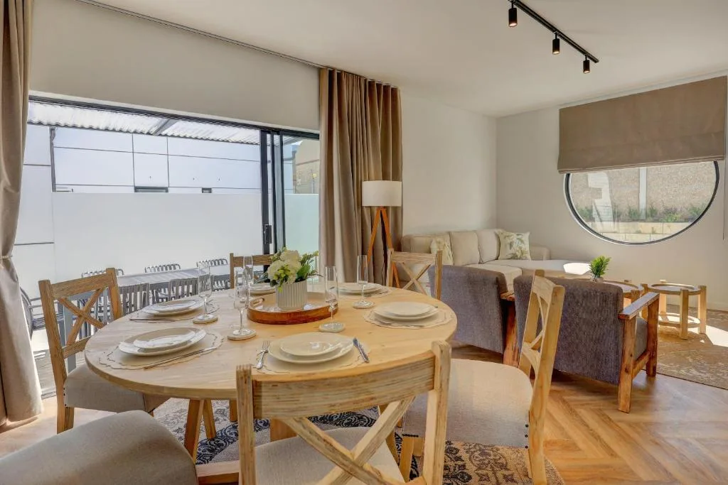 Light wooden dining table set for six with lounge area visible beyond