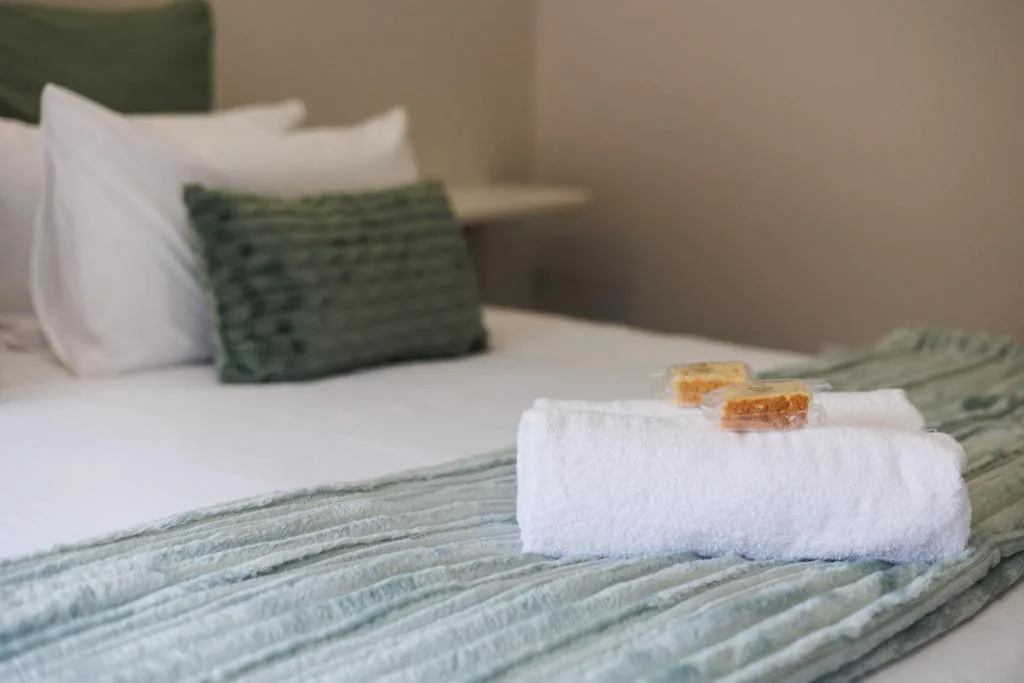 Neatly made bed with white pillows, green cushion, and rolled white towels with wooden accents