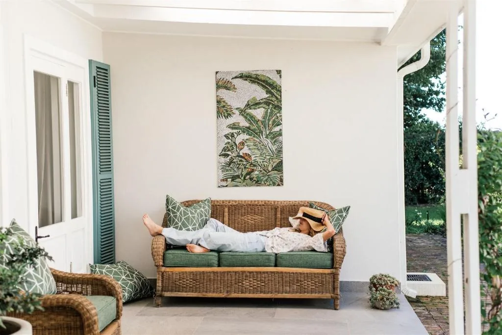 Covered patio with wicker sofa, green cushions, botanical artwork, and garden views