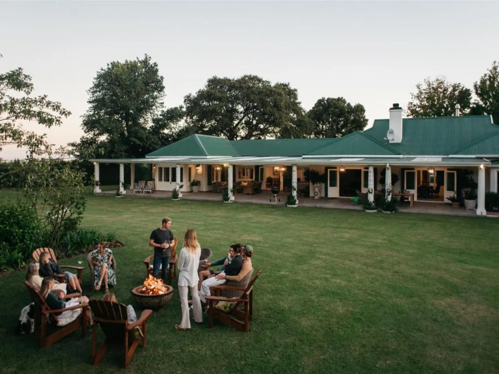 Large colonial-style home with green roof and white columns, guests gathered around fire pit on lawn