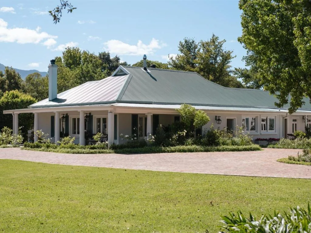 White Cape Dutch style villa with metal roof and wraparound veranda, green lawn and gardens