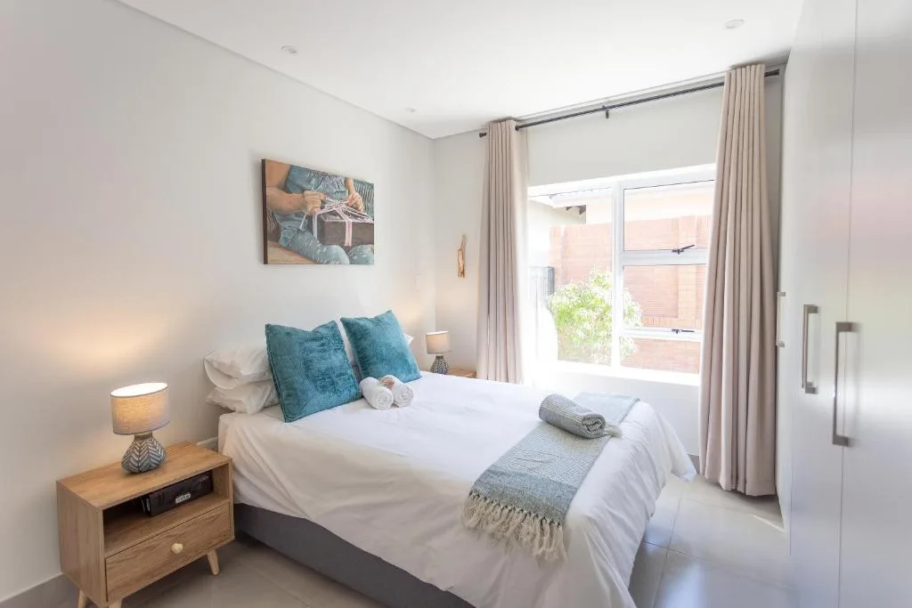 Bright bedroom with white bedding, teal pillows, wooden nightstand, and garden view